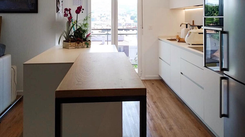 Cocina doméstica moderna con encimera de madera, muebles blancos y suelo laminado, iluminada por una gran ventana al exterior.