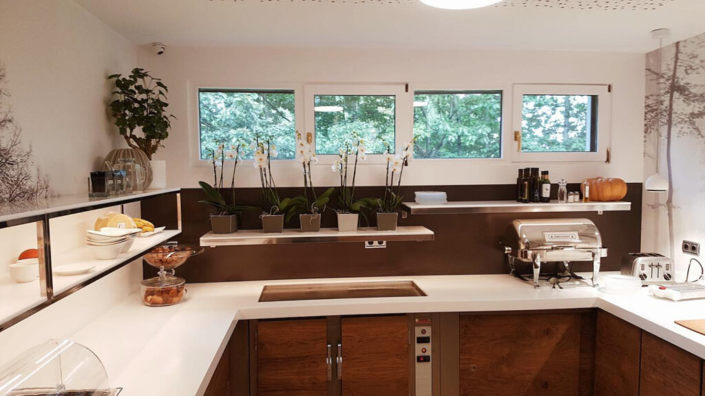 Zona de buffet en cocina moderna con encimera blanca, flores en macetas, bandejas con fruta, bollería y calentador de inox para comidas.